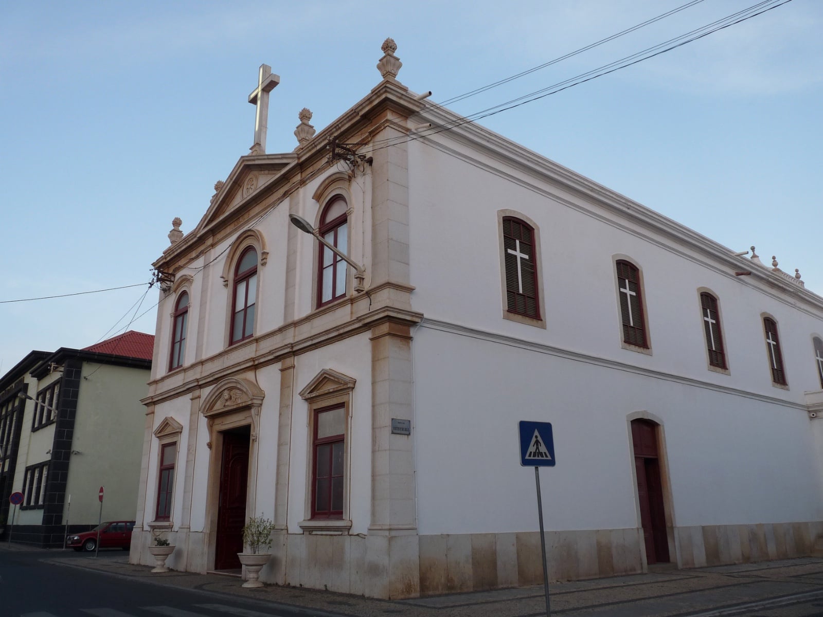 Igreja católica de fachada branca vista da esquina da rua, com cruz de pedra no frontão e janelas de caixilharia vermelha